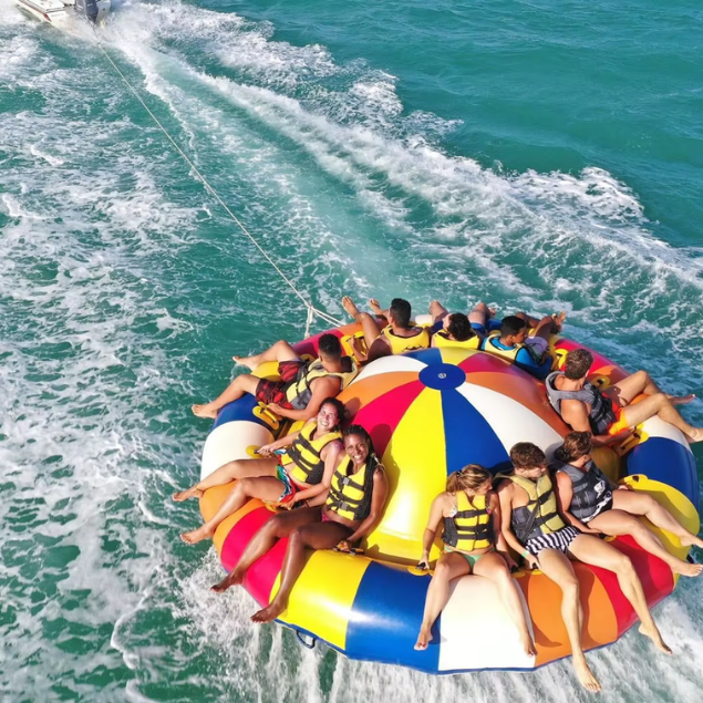 Group of smiling people riding a colorful UFO-shaped towable inflatable across the turquoise water during